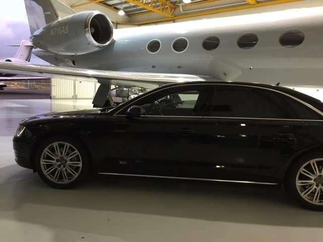 A black Audi luxury chauffeur car parked at an airport terminal, with a chauffeur ready for corporate transfers